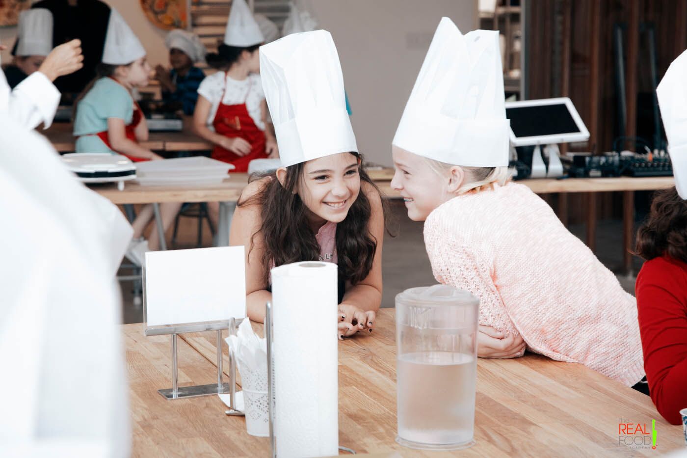 Two young chefs whispering and giggling to each other at a The Real Food Academy cooking camp.
